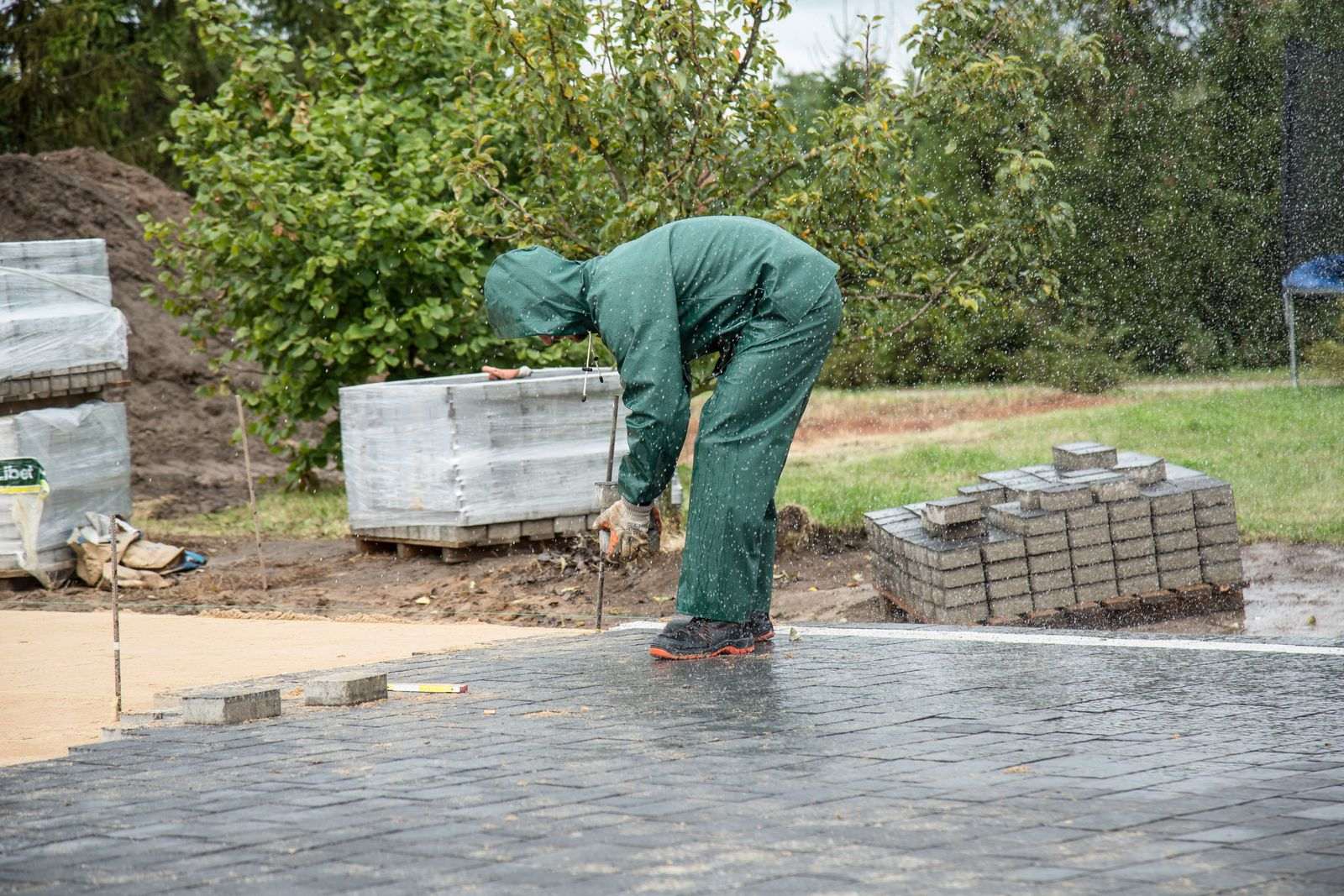 A worker in a green rain suit laying grey paving stones on a wet patio